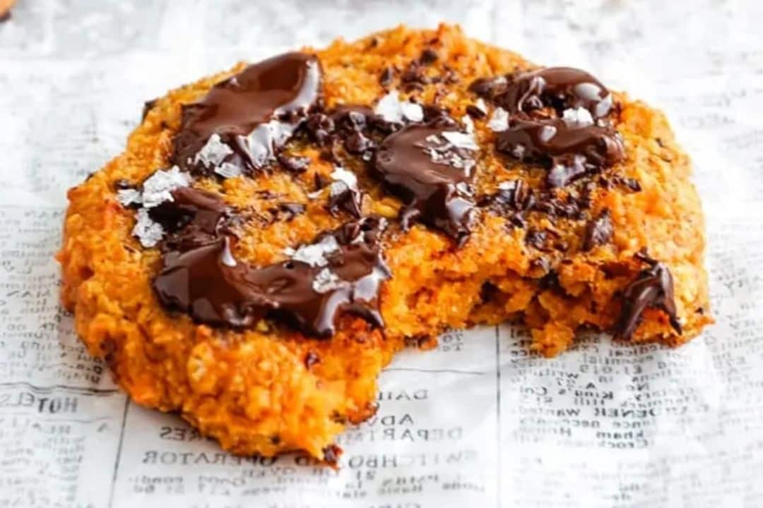 A large chocolate chip cookie with sea salt flakes, partially eaten, sits on a sheet of newspaper—next to notes on sweet potato recipes.