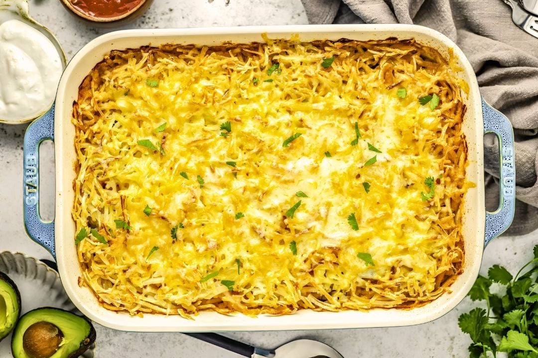 Baked casserole topped with melted shredded cheese, garnished with chopped herbs, in a white dish surrounded by avocado, cilantro, sour cream, and a brown sauce—perfect for those seeking cozy potato side dishes and hearty fall dinner inspiration.