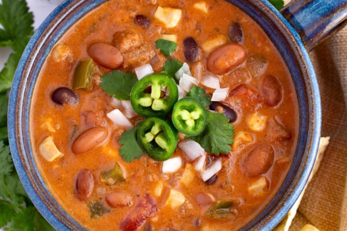 A bowl of chili soup with beans, cheese, diced onions, cilantro, and sliced jalapeños on top, served in a blue dish—perfect for anyone looking to add bold flavors to their fall recipes or expand their collection of hearty soup recipes.