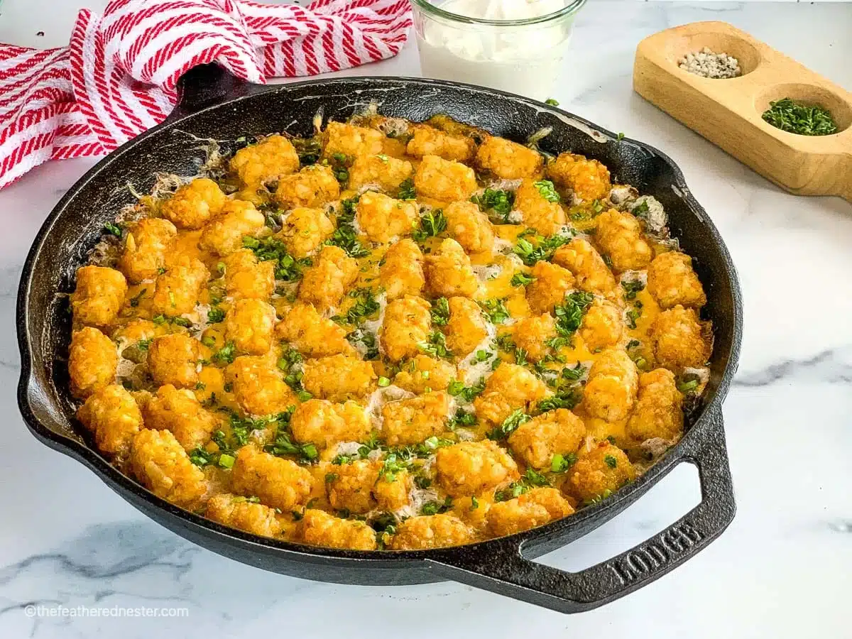 A cast iron skillet filled with baked tater tot casserole, one of the best easy casseroles, is topped with golden tater tots and green herbs. It sits on a marble surface beside seasonings and a bowl of sour cream, perfect inspiration for your next round of dinner recipes.