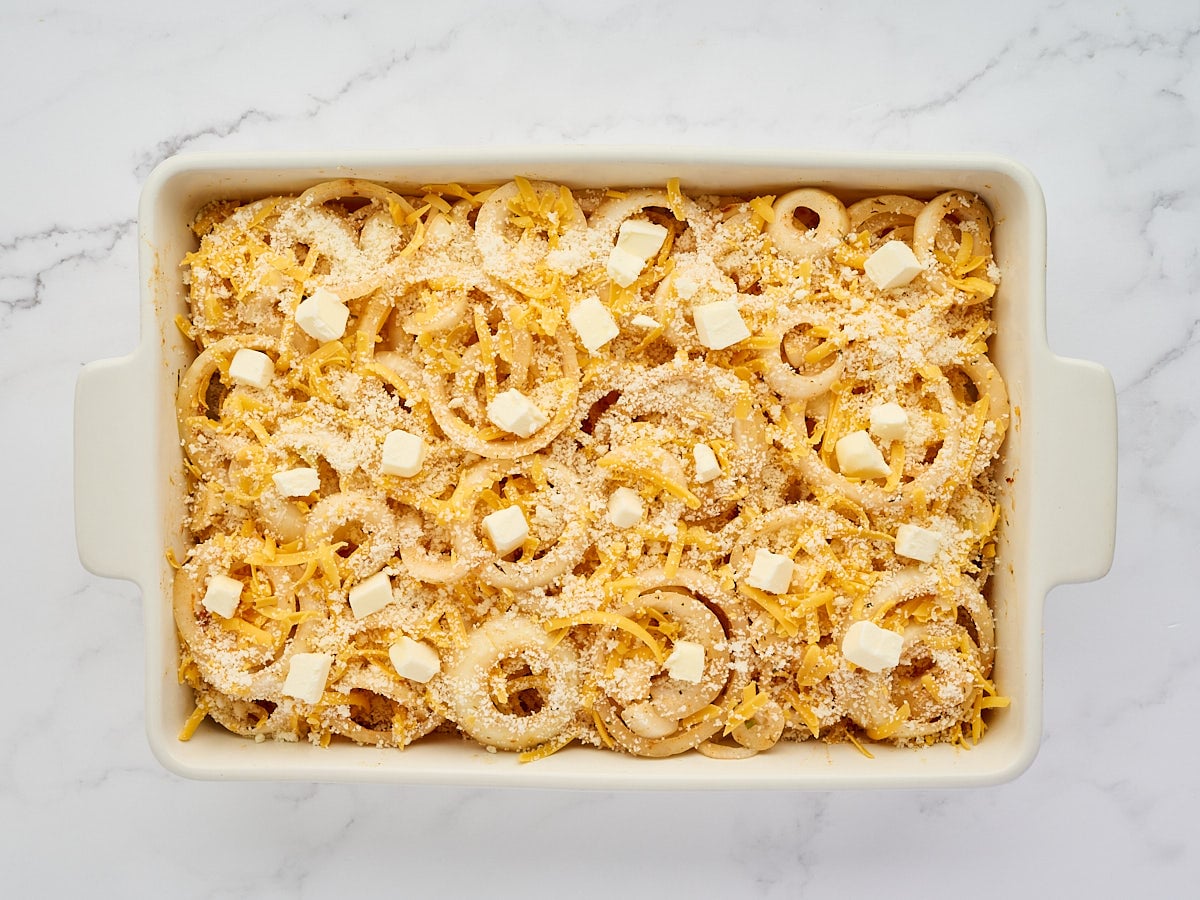 A white baking dish filled with raw sliced onions, shredded cheese, breadcrumbs, and small cubes of butter on a marble surface.