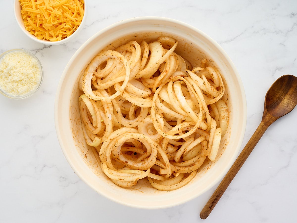 A bowl of sliced onions seasoned with spices sits next to a wooden spoon, with small bowls of shredded cheddar and grated Parmesan cheese nearby on a marble surface.