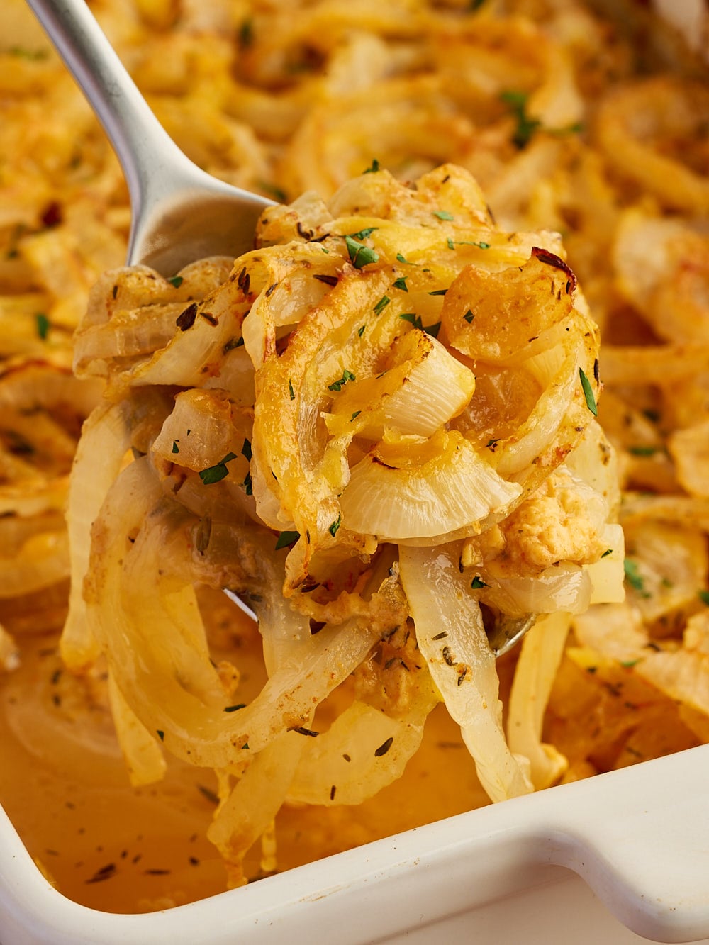 A serving spoon lifts cooked sliced onions with herbs from a white baking dish filled with more onions and broth.