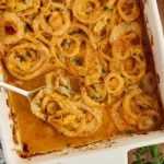 Baked casserole with sliced onions in a creamy, orange-colored sauce, garnished with chopped herbs, in a white dish with a serving spoon.