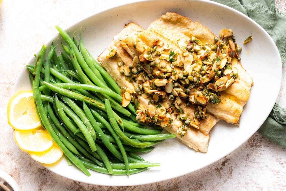 A plate with sautéed fish topped with sliced almonds and herbs, served with steamed green beans and lemon slices on the side—this dish is perfect for anyone looking for easy fish dishes to add to their seafood recipes collection.