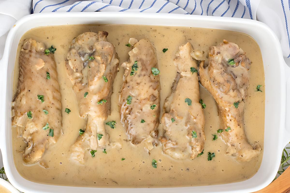 Four cooked chicken legs in a creamy gravy sauce, garnished with chopped herbs and arranged in a white baking dish—a classic comfort food perfect for Sunday suppers and home cooking.