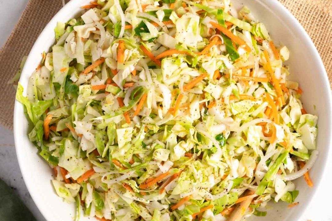 A white bowl filled with shredded cabbage, carrots, and seasonings sits on a beige placemat, making it a fresh and vibrant addition to your collection of delicious cabbage recipes.