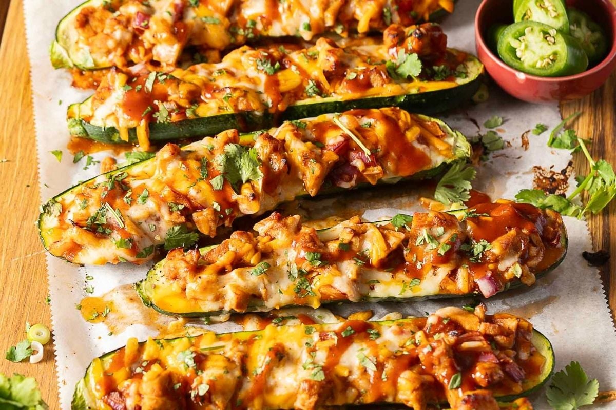 Zucchini boats filled with melted cheese, chicken, sauce, and herbs sit on parchment paper next to a small bowl of sliced jalapeños—perfect for those searching for new dinner recipes or looking to add to their collection of saved recipes.