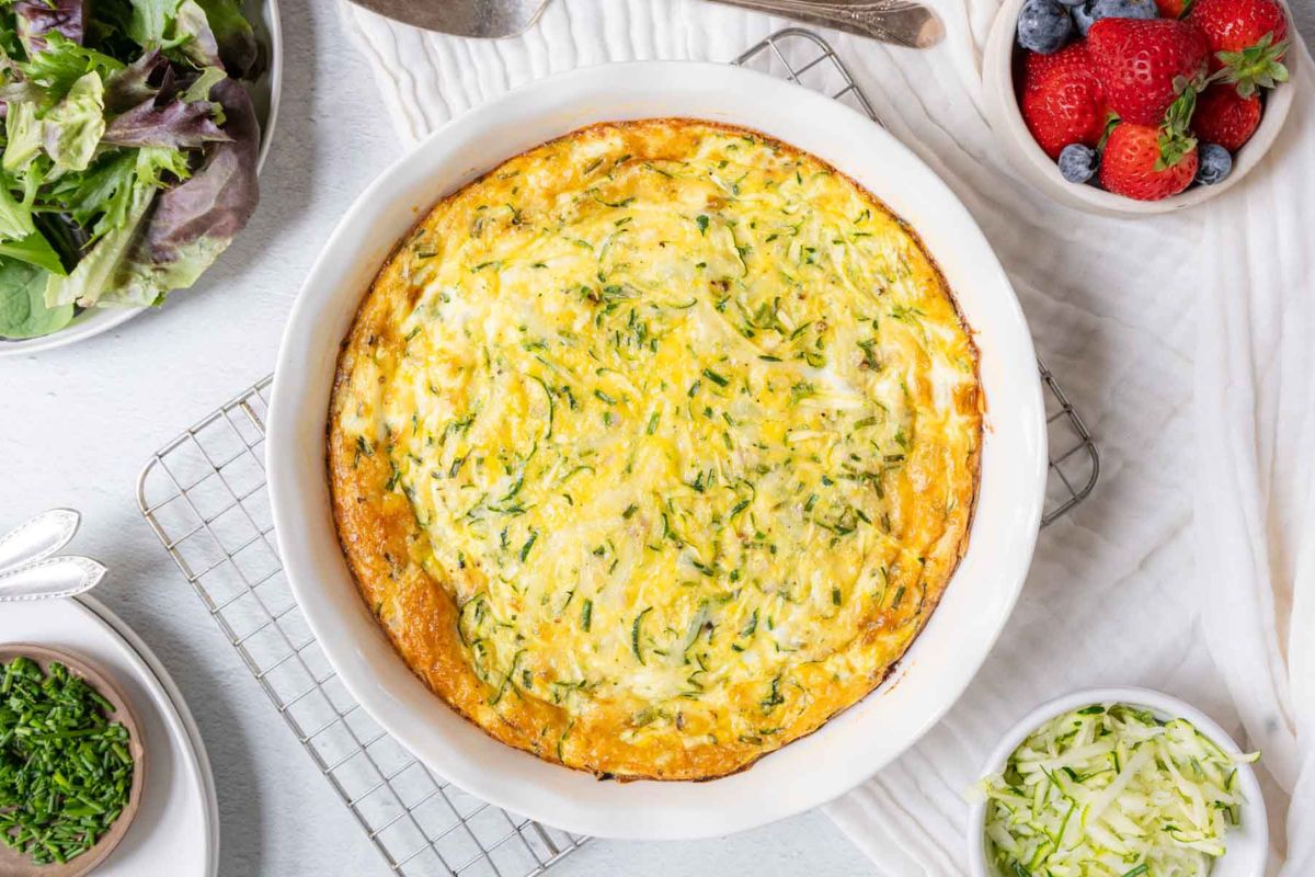 Baked zucchini frittata in a white dish on a cooling rack, surrounded by fresh salad, berries, and bowls of chopped herbs and grated zucchini—perfect for those who love savory breakfasts or are searching for new savory breakfast recipes.
