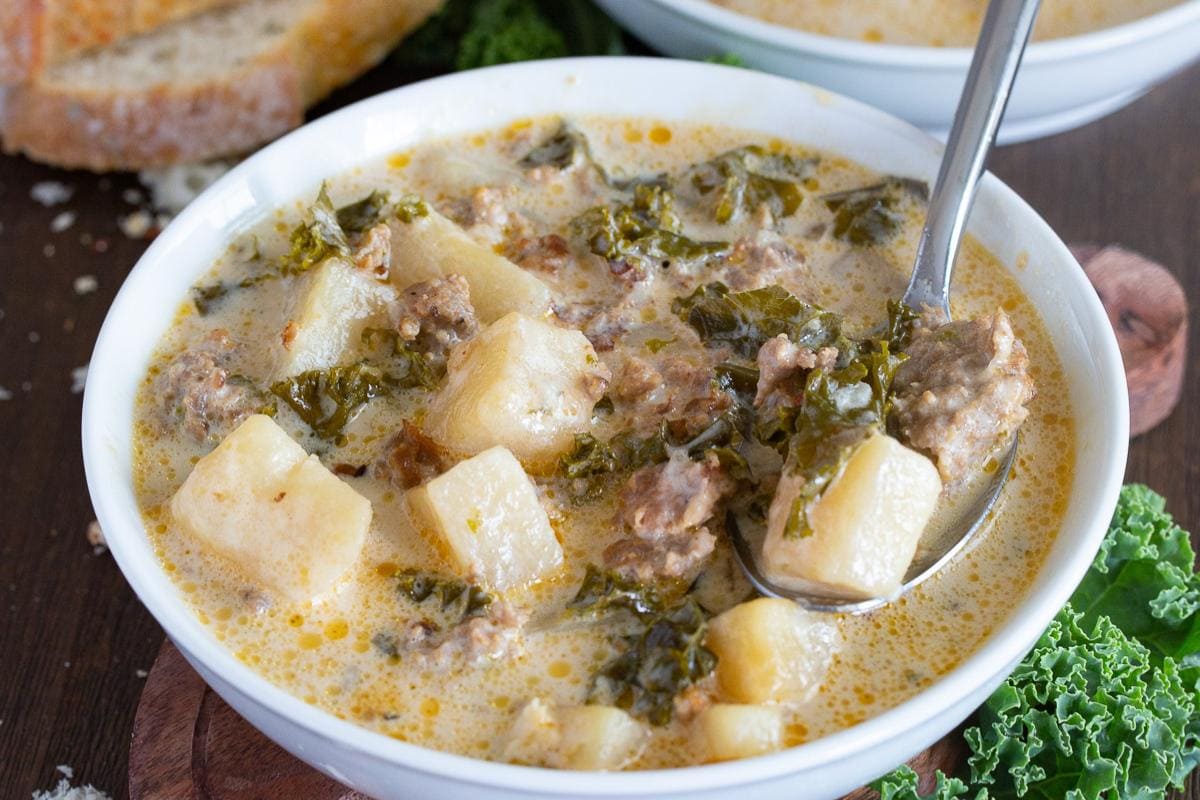 A bowl of creamy soup containing sausage, potatoes, and kale, with a spoon resting inside the bowl. Slices of bread are visible in the background—perfect for anyone seeking comforting fall recipes or delicious creamy soup recipes to warm up chilly evenings.