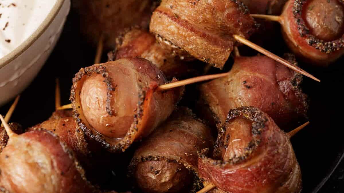 Bacon-wrapped sausages secured with toothpicks, served with a side of white dipping sauce as savory potato side dishes.