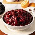 A bowl of cranberry sauce on a plate, with oranges and blackberries in the background.