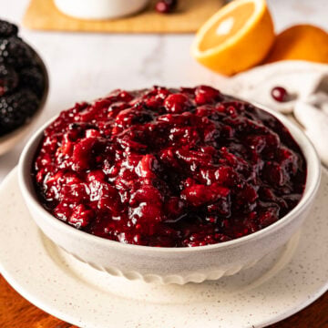A bowl of cranberry sauce on a plate, with oranges and blackberries in the background.