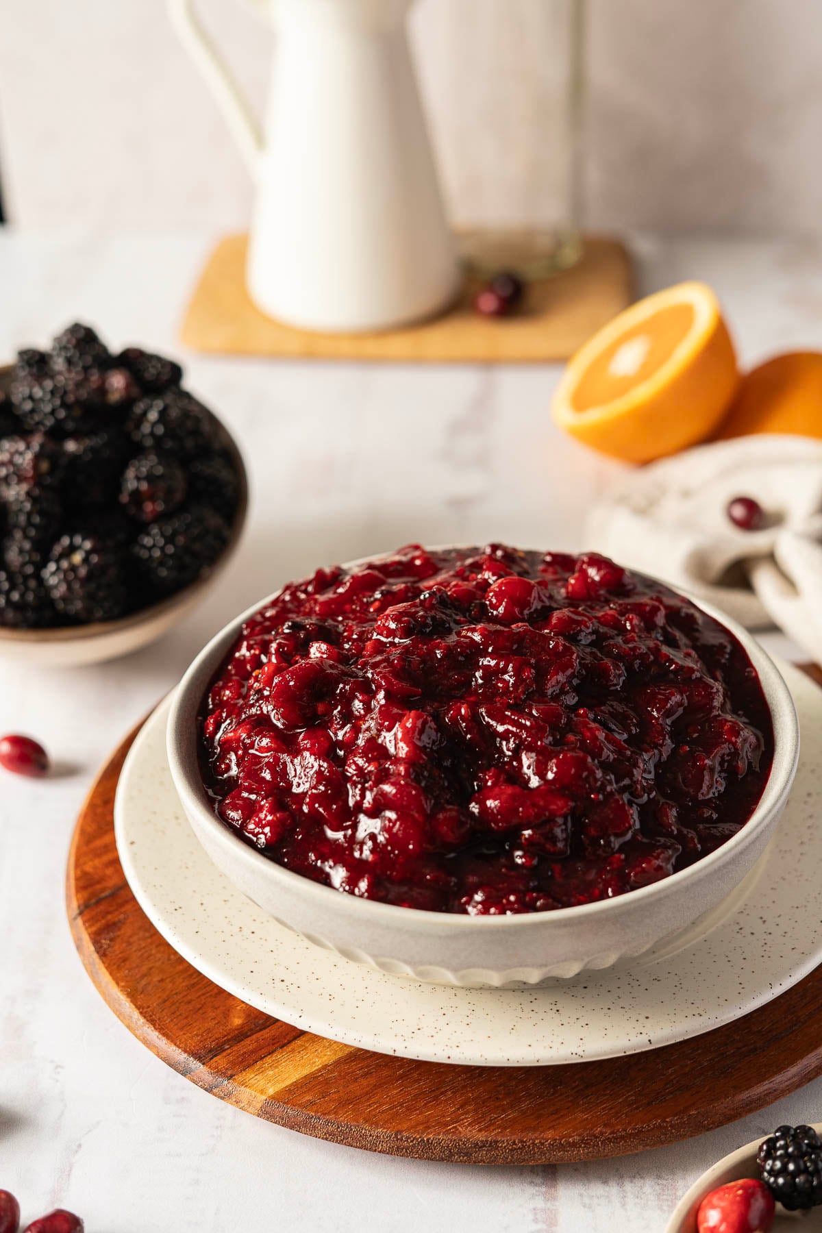 A bowl of dark red cranberry sauce on a wooden board, surrounded by blackberries and orange slices.