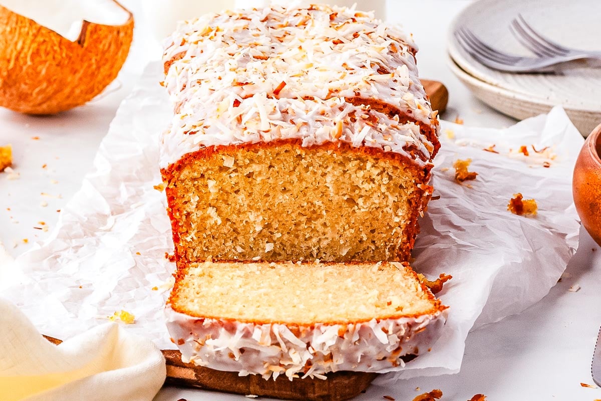 A loaf of coconut bread with white icing and shredded coconut on top sits on parchment paper, with one slice cut and lying in front.