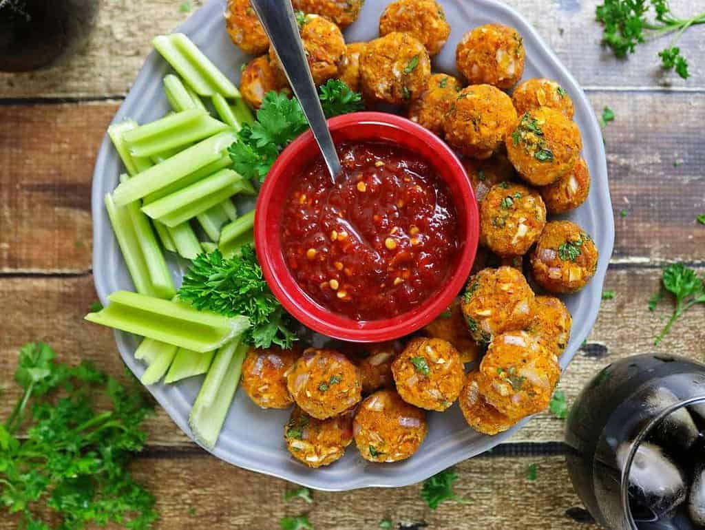 A plate of butternut squash fritters served with celery sticks and a bowl of red dipping sauce.