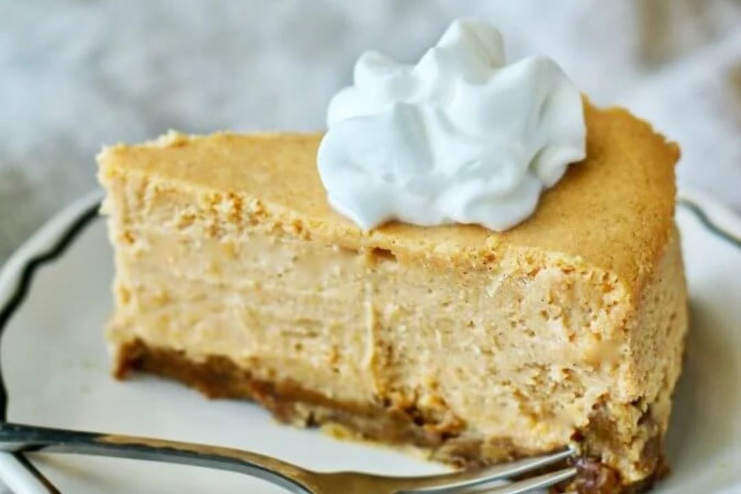 A slice of butternut squash cheesecake topped with a dollop of whipped cream on a plate with a fork.