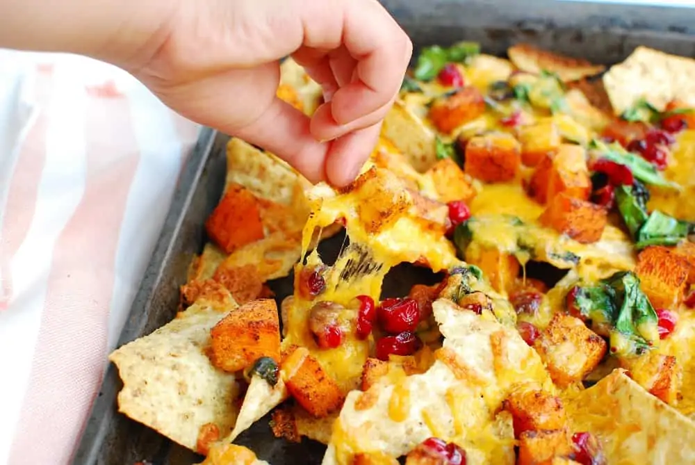 A hand lifts a tortilla chip covered in melted cheese, roasted squash, and cranberries from a baking sheet, showcasing simple butternut squash recipes.