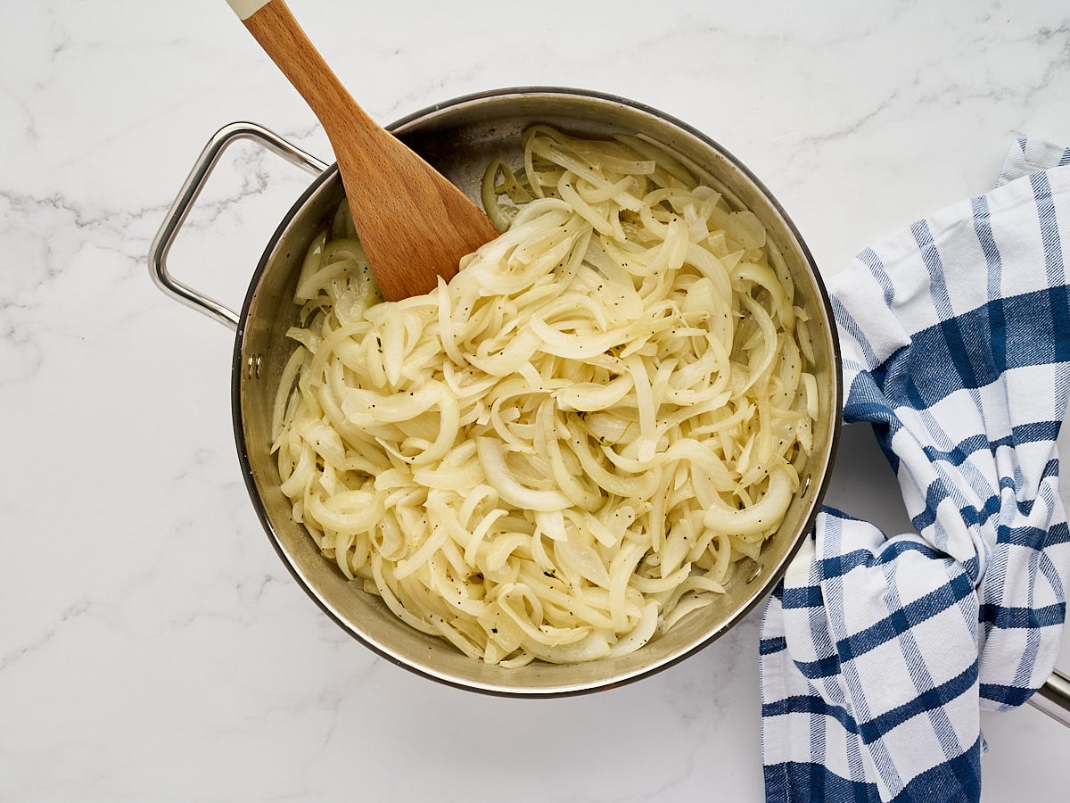 Sliced onions cooking in a stainless steel pan with a wooden spoon and a blue and white checkered kitchen towel.