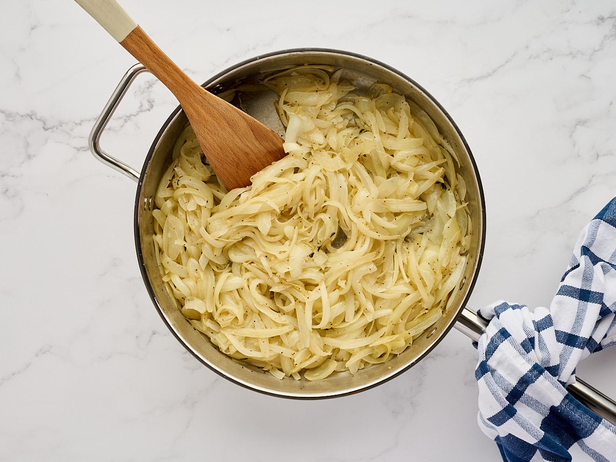 Sliced onions cooking in a stainless steel pan with a wooden spoon.