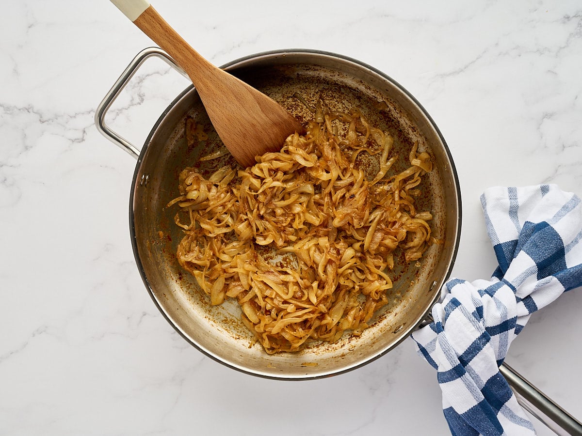 A wooden spoon stirs caramelized onions in a stainless steel skillet.