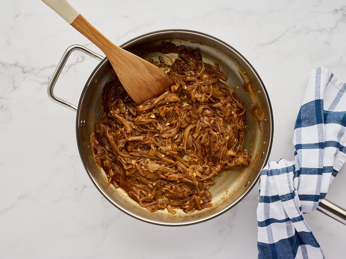 A metal skillet containing caramelized onions being stirred with a wooden spoon, next to a blue and white checkered kitchen towel.