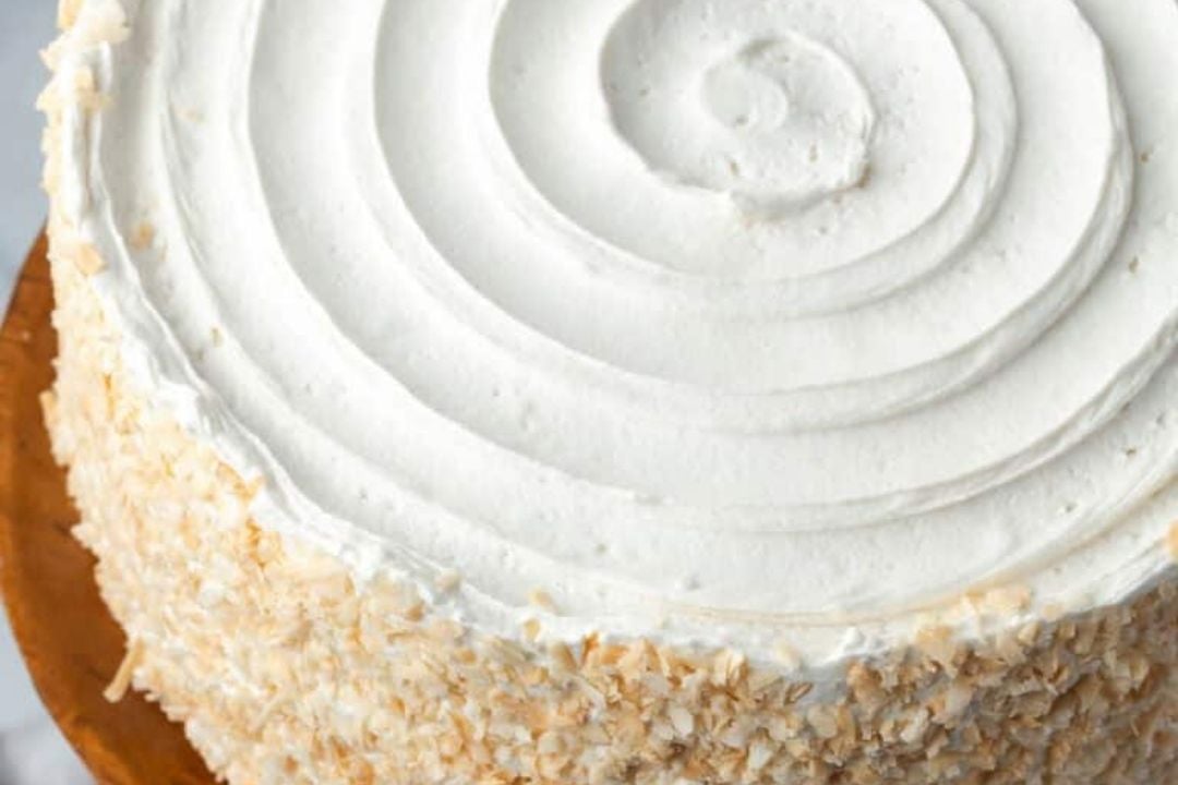 Round cake with smooth white frosting and toasted coconut flakes on the sides, displayed on a wooden cake stand.