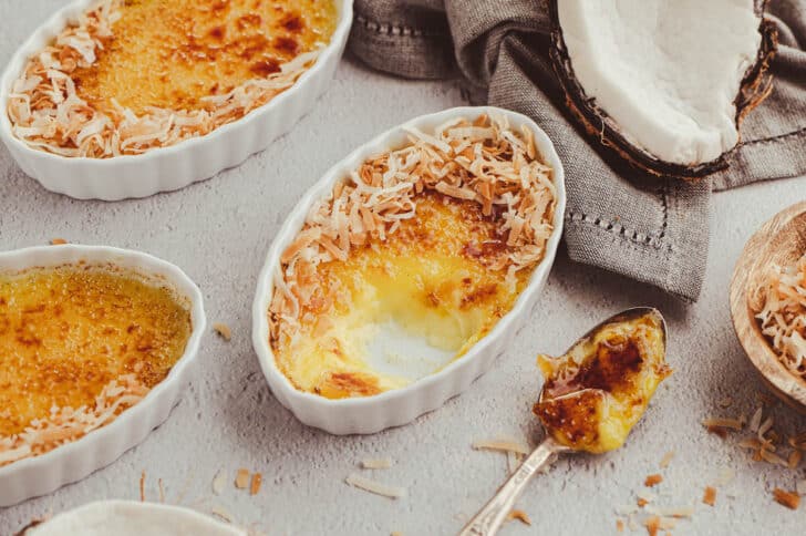 Oval ramekins with crème brûlée topped with toasted coconut, one partially eaten, on a light surface with a spoon and coconut shell nearby.