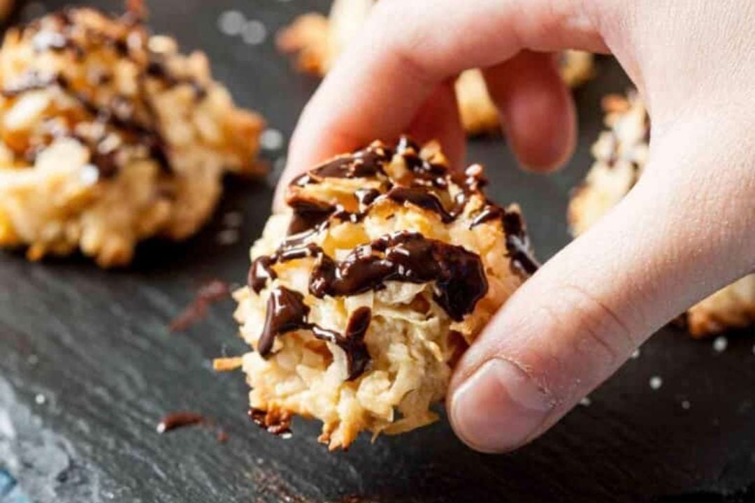 A hand picks up a coconut macaroon drizzled with chocolate from a slate surface, with other similar cookies in the background.