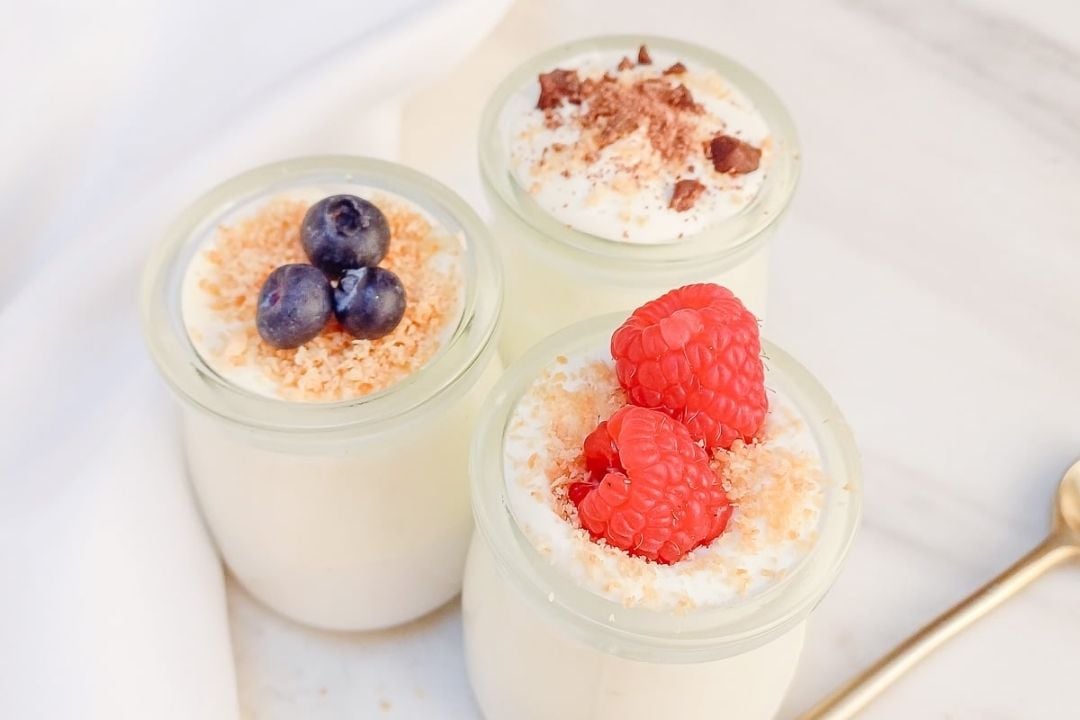Three glass jars of creamy dessert topped with berries, toasted crumbs, and chocolate shavings, arranged on a white surface with a gold spoon nearby.