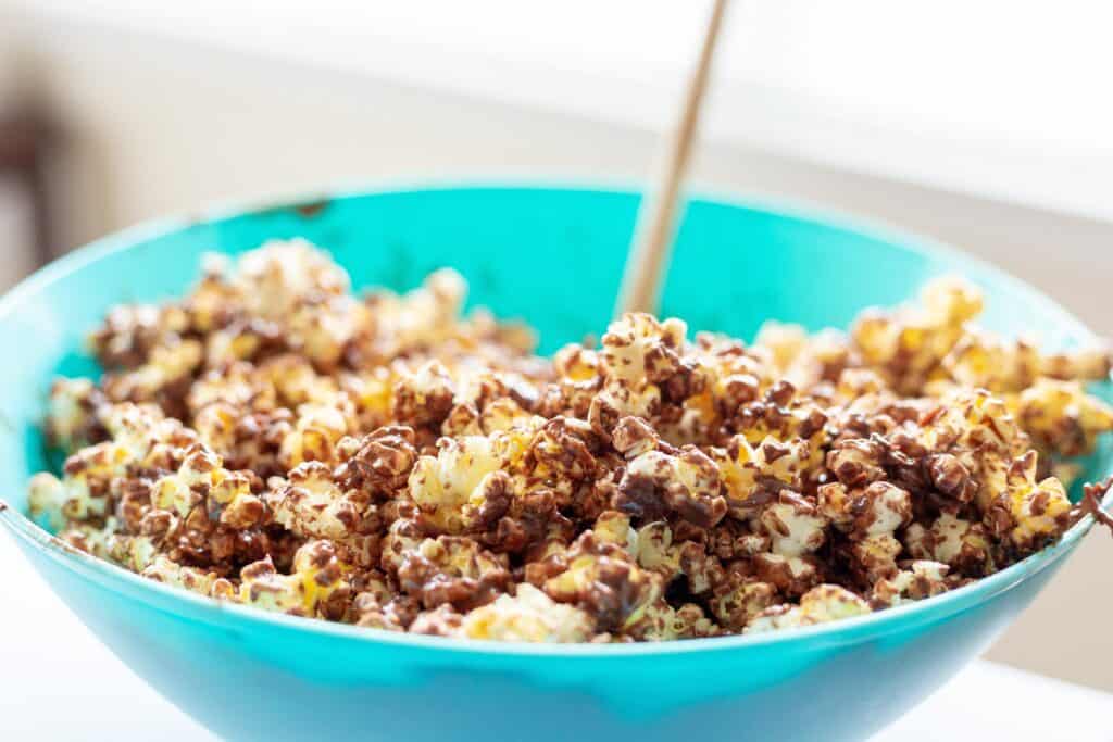 A blue bowl filled with popcorn coated in a chocolate drizzle, with a wooden spoon partially visible in the background.