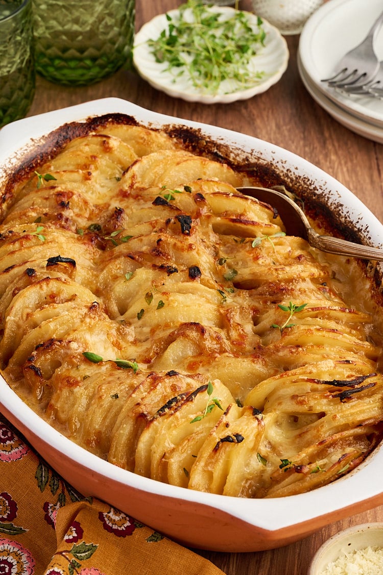 A scalloped potato gratin in an oval baking dish, topped with fresh herbs and a serving spoon.