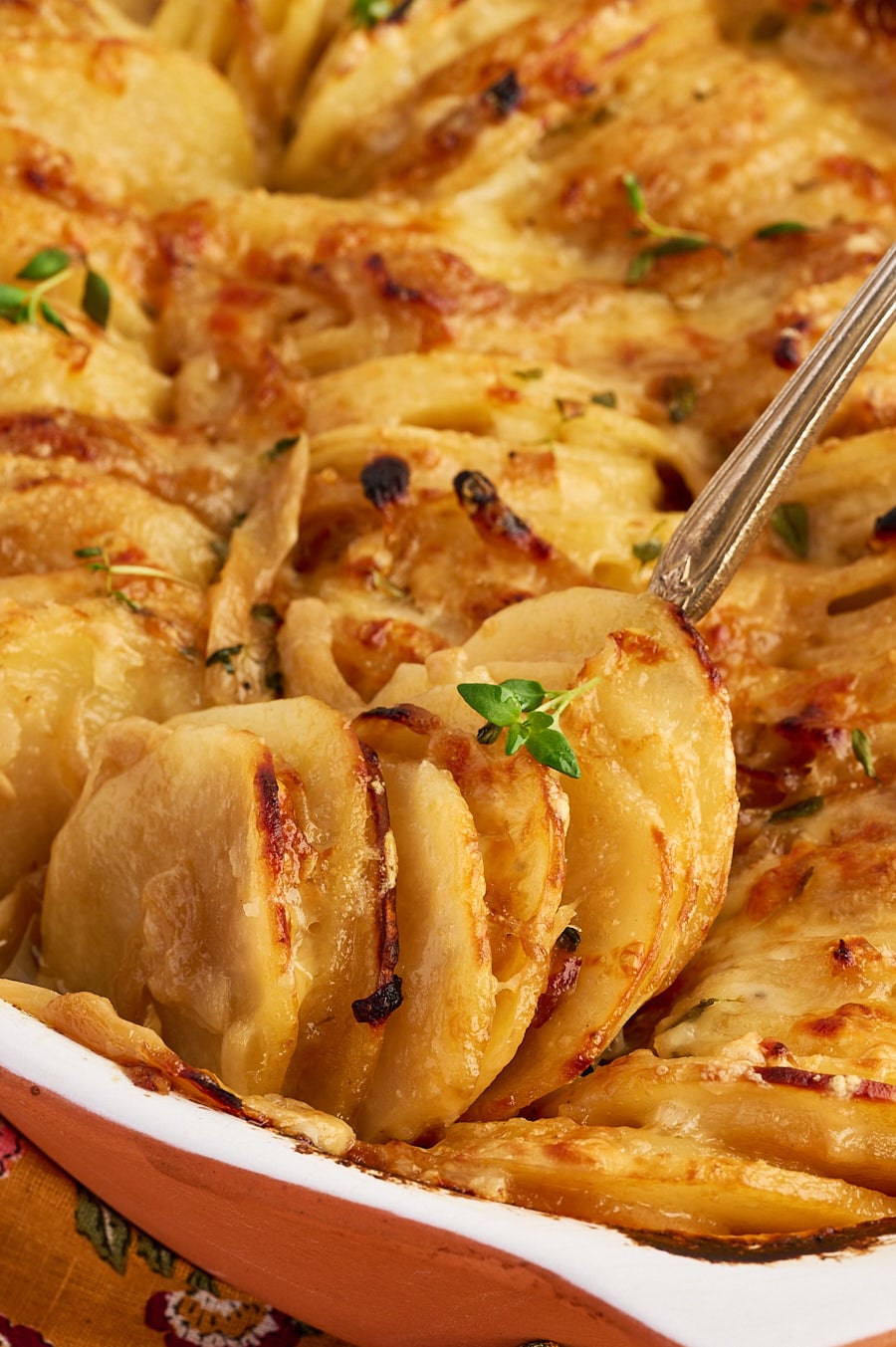 A serving spoon lifts a portion of scalloped potatoes from a baking dish, garnished with fresh thyme.