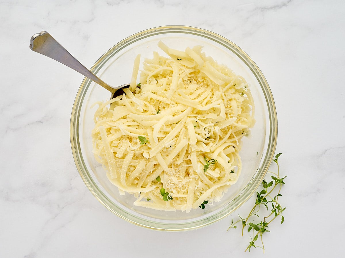 A glass bowl of shredded cheese with fresh herbs and a silver spoon.