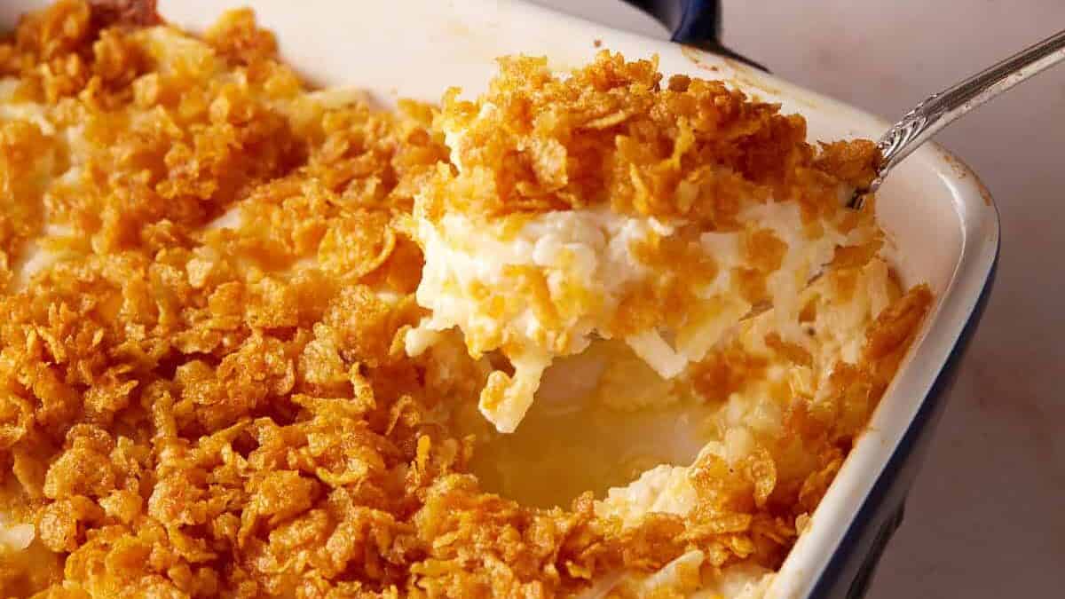 A serving spoon lifts a portion of cheesy potato casserole topped with cornflakes from a baking dish, a classic among potato side dishes.