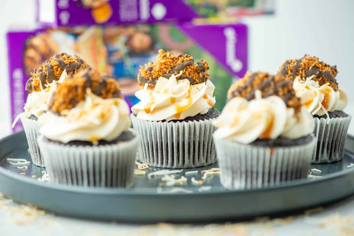 Four chocolate cupcakes with white frosting and caramel cookie topping sit on a round tray, with purple cookie boxes in the background.