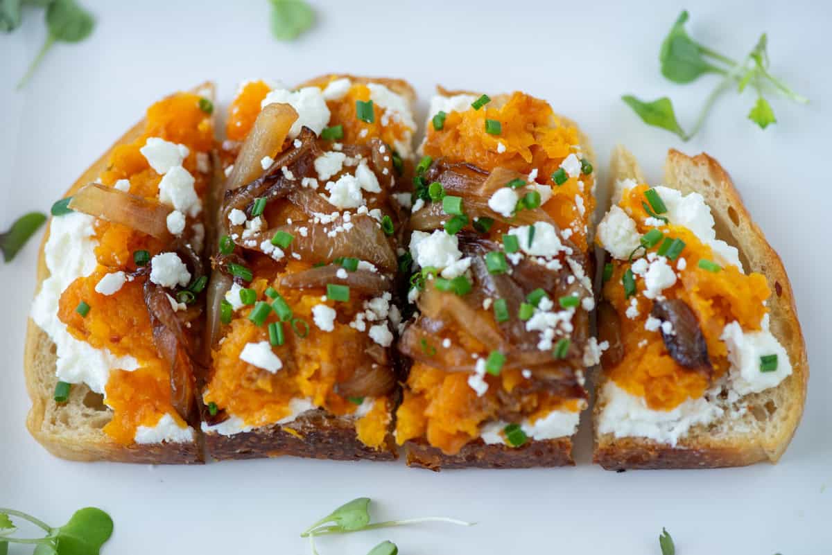 Four slices of toasted baguette topped with mashed butternut squash, caramelized onions, crumbled goat cheese, and chives.