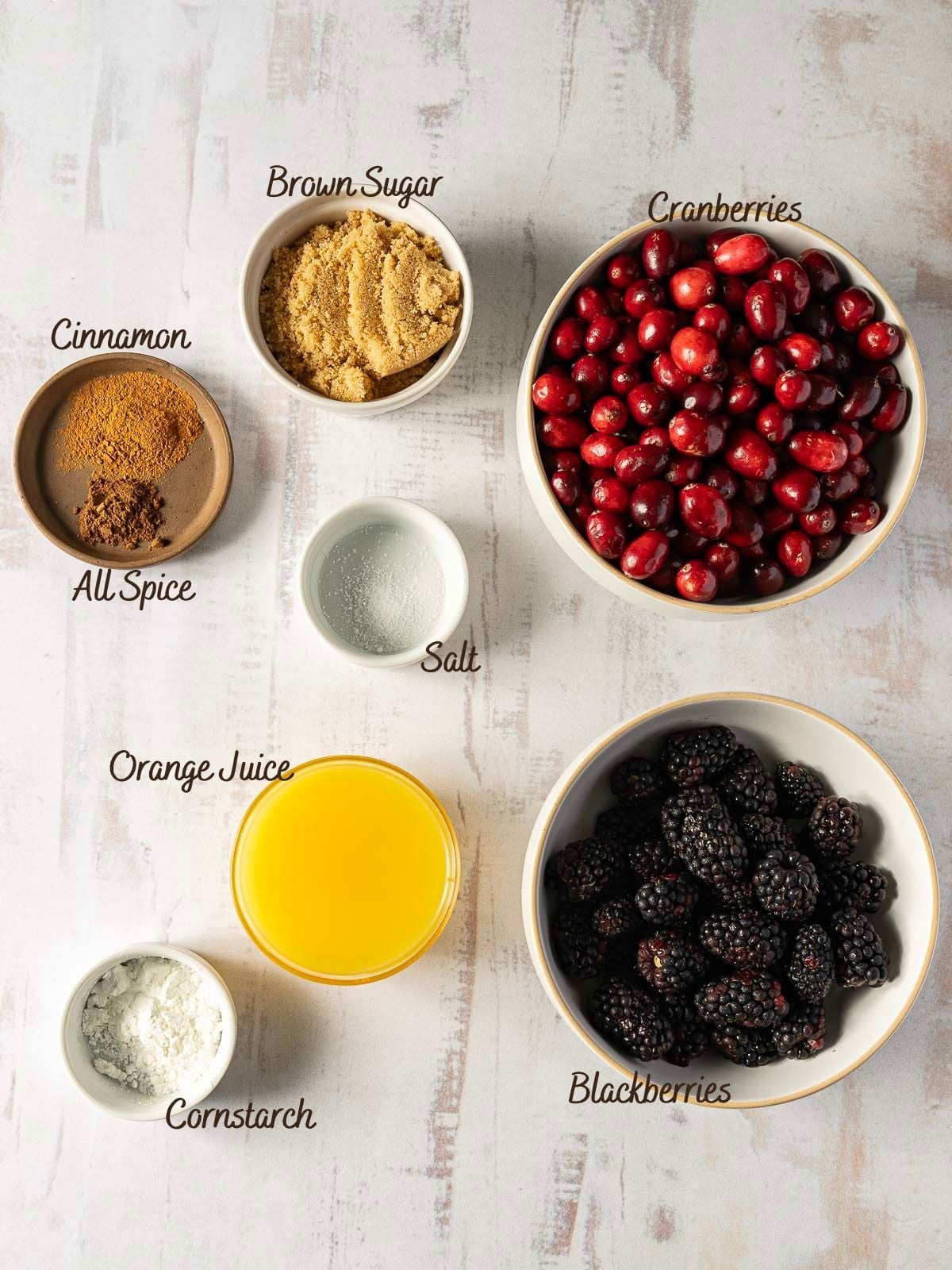 Ingredients for a blackberry cranberry sauce recipe arranged in bowls and ramekins, labeled: brown sugar, cranberries, cinnamon, allspice, salt, orange juice, cornstarch, and blackberries.