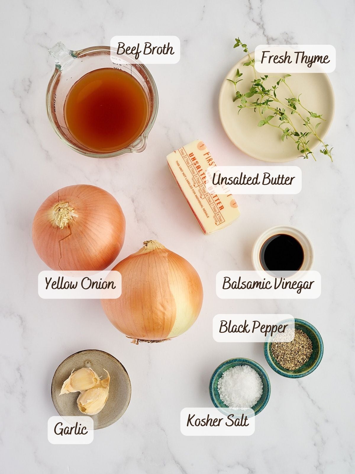 Ingredients for caramelized onions on a marble surface, including beef broth, fresh thyme, unsalted butter, yellow onions, balsamic vinegar, black pepper, kosher salt, and garlic.