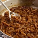 A metal spoon lifts caramelized onions from a stainless steel pan.