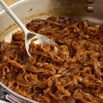 A metal spoon lifts caramelized onions from a stainless steel pan.