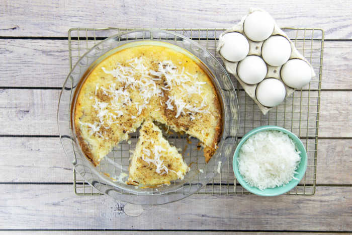 A coconut pie with one slice missing sits on a cooling rack next to a bowl of shredded coconut and a carton of eggs.