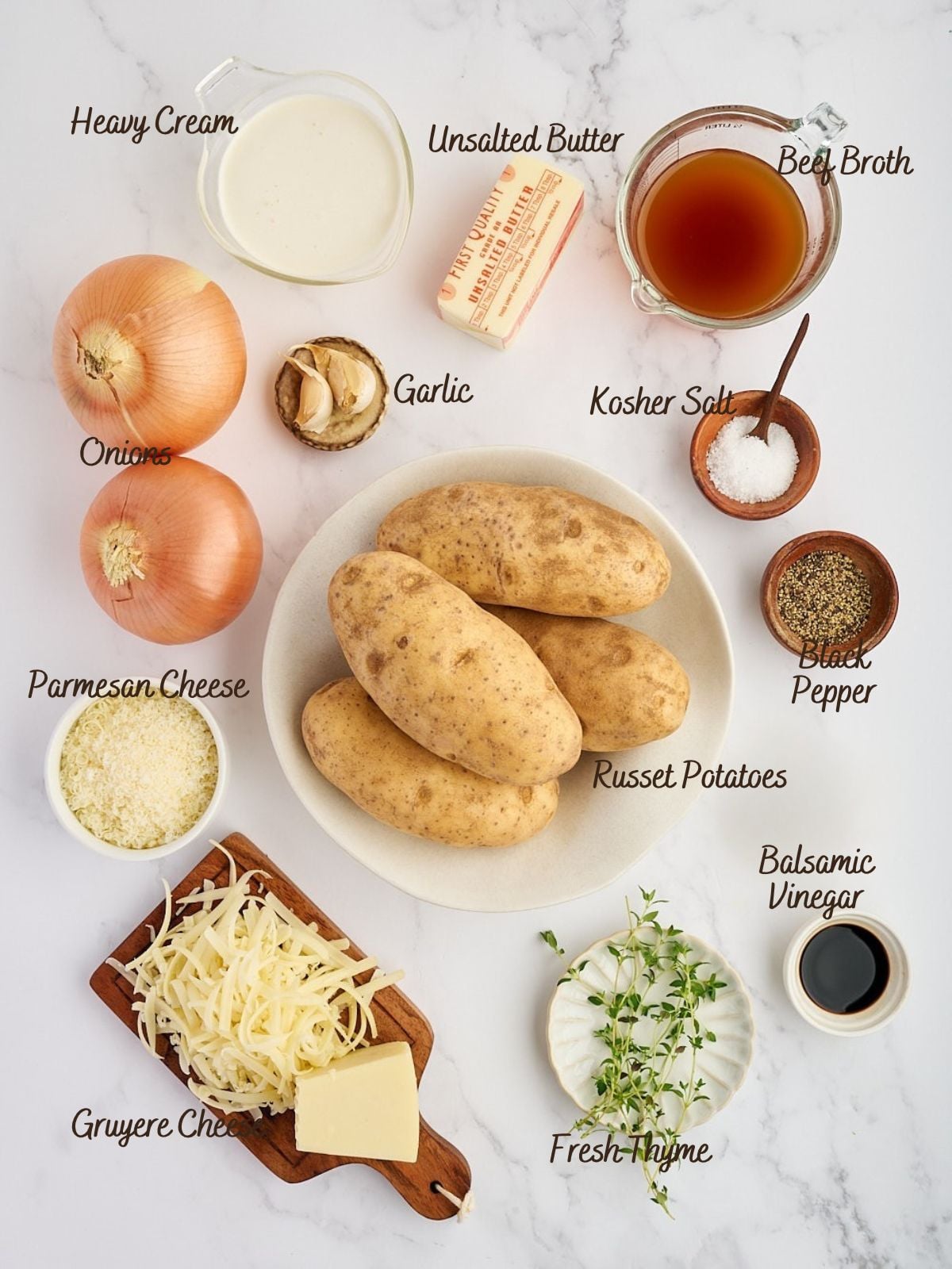 Ingredients for French onion potatoes: heavy cream, butter, beef broth, onions, garlic, salt, pepper, parmesan, russet potatoes, gruyere, balsamic vinegar, and fresh thyme on a white surface with labels.