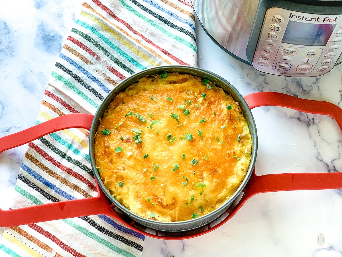 A round potato side dish with a melted cheese crust, topped with herbs, in a red silicone sling next to an Instant Pot.