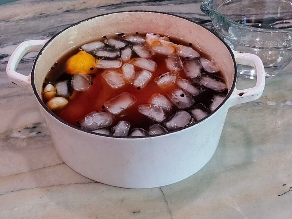 A white pot containing a dark liquid with ice cubes, a whole lemon, and garlic cloves for a slow cooker turkey breast.