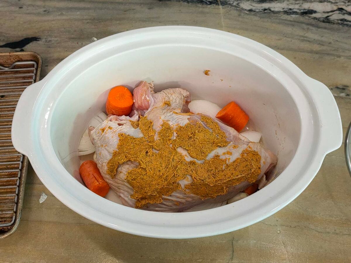 A seasoned slow cooker turkey breast surrounded by carrot chunks and onion slices.