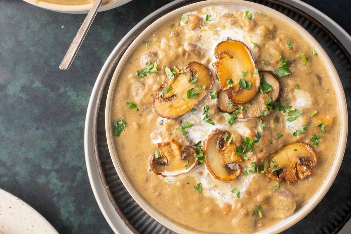 A bowl of comforting creamy mushroom soup garnished with sliced sautéed mushrooms and fresh herbs.