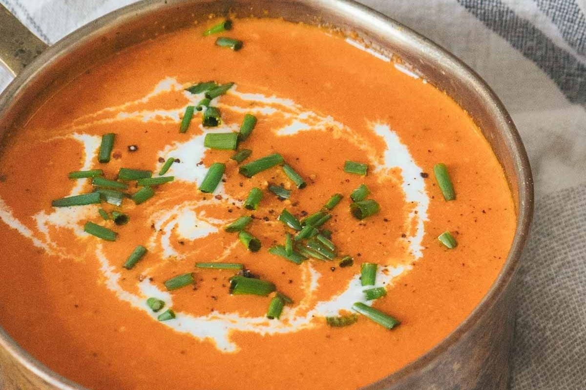 A metal bowl of comforting creamy tomato soup swirled with cream and topped with chopped chives.