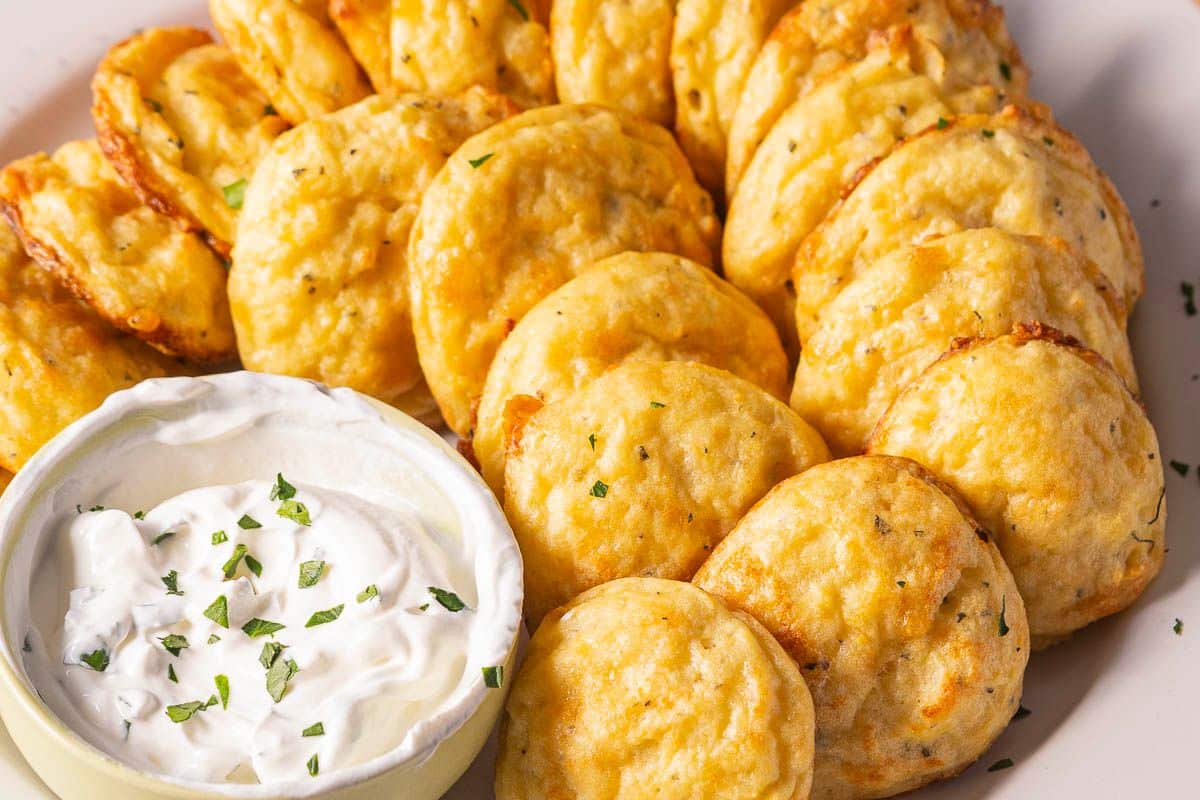 A plate of round, golden-brown cheese fritters, a classic among potato side dishes, served with a bowl of sour cream topped with herbs.